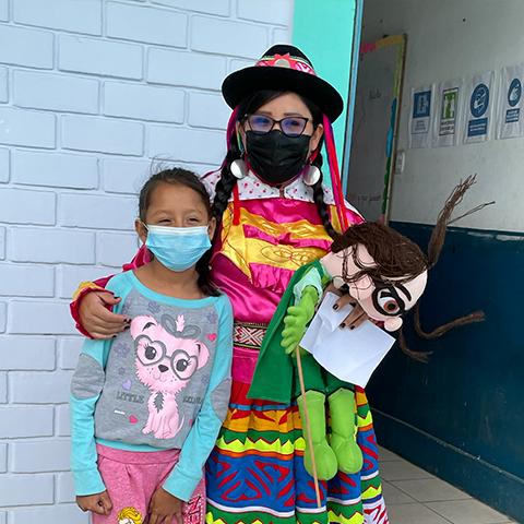 Niña posa junto a una mujer con traje tradicional y un personaje educativo.