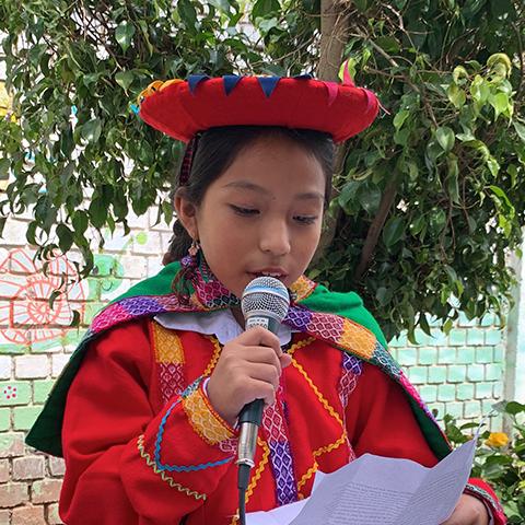 Niña con traje tradicional sostiene un micrófono durante una actividad escolar.